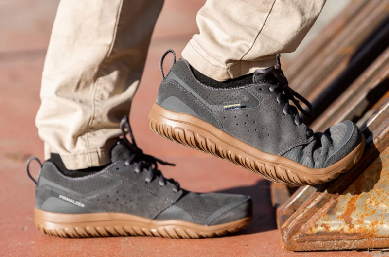 Load image into Gallery viewer, A person wearing dark gray suede and mesh outdoor shoes with tan soles, standing on a rusty metal structure. The brand name, Lems is visible on the side of the shoe.