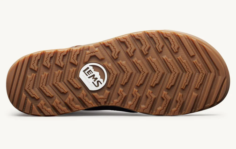Load image into Gallery viewer, The textured brown rubber outsole of a shoe, featuring a zigzag tread pattern and a circular white and black Lems logo in the center, shown against a clean off-white background.
