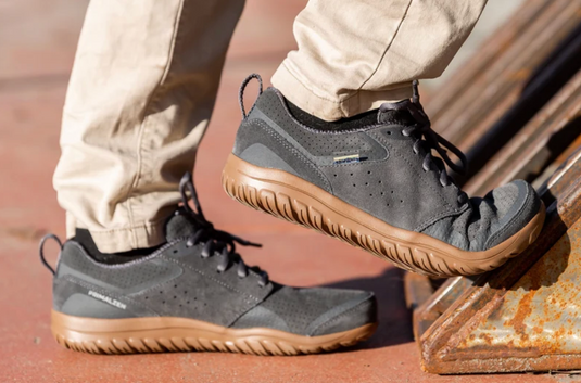 A person wearing dark gray suede and mesh outdoor shoes with tan soles, standing on a rusty metal structure. The brand name, Lems is visible on the side of the shoe.